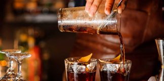 Bartender pouring cocktails into two glasses with lemon garnishes.