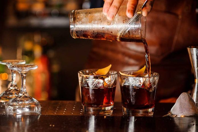 Bartender pouring cocktails into two glasses with lemon garnishes.