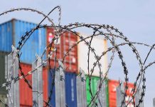 Barbed wire fence with shipping containers in background.