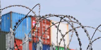 Barbed wire fence with shipping containers in background.