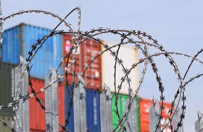 Barbed wire fence with shipping containers in background.