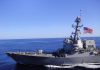 Warship sailing with American flag in open sea.