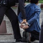 Person kneeling handcuffed by police officers outside